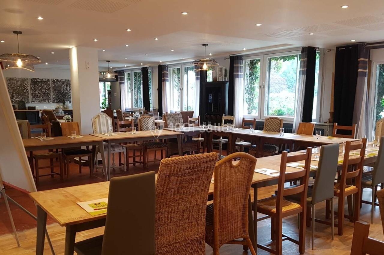 Salle lumineuse aménagée avec de longues tables en bois et des chaises variées, entourée de grandes fenêtres.