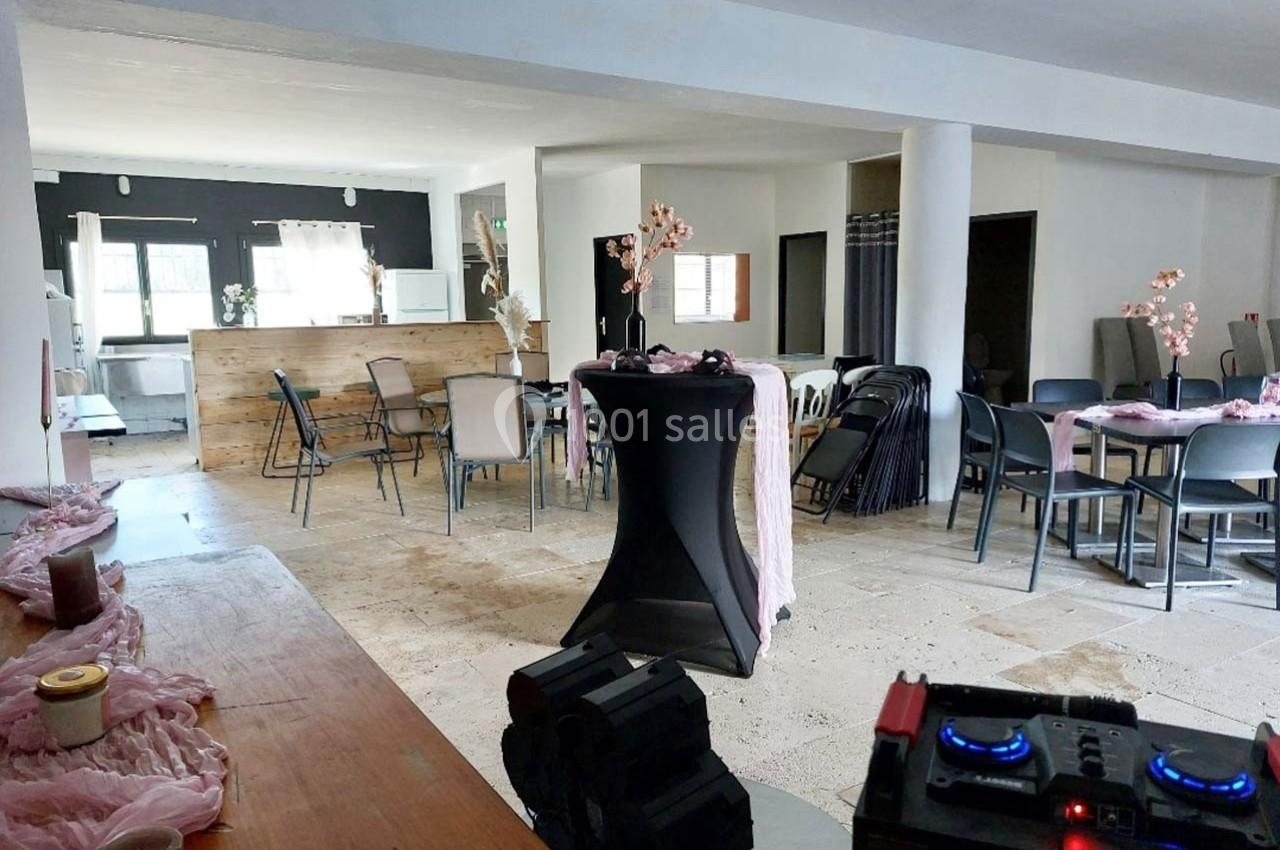 Salle lumineuse aménagée pour un événement, avec tables décorées, chaises pliantes et un espace bar en arrière-plan.