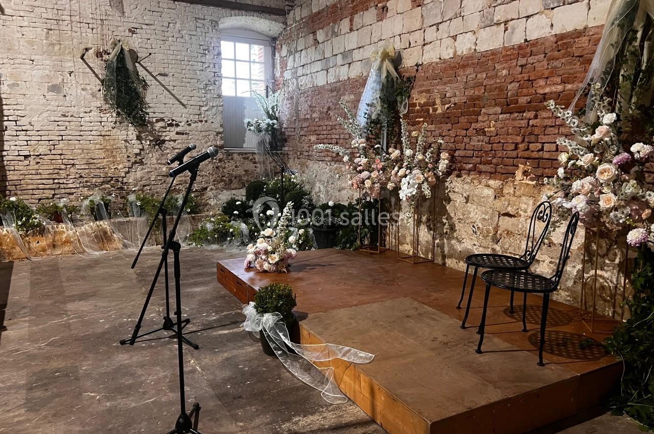 Scène décorée de fleurs dans une salle en briques, avec un micro sur pied et une chaise en métal.