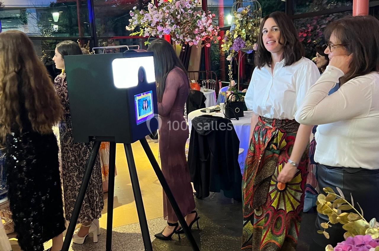 Des personnes posent près d'une borne photo dans un espace intérieur décoré de fleurs et éclairé par des lumières colorées.