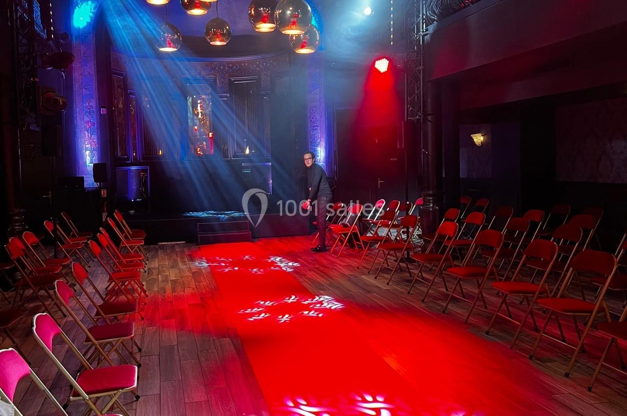 Salle de spectacle avec un tapis rouge central, des chaises alignées et des éclairages colorés.