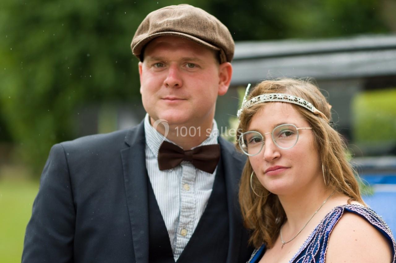 Un homme en costume avec un nœud papillon et une casquette pose à côté d'une femme portant une robe et un bandeau.