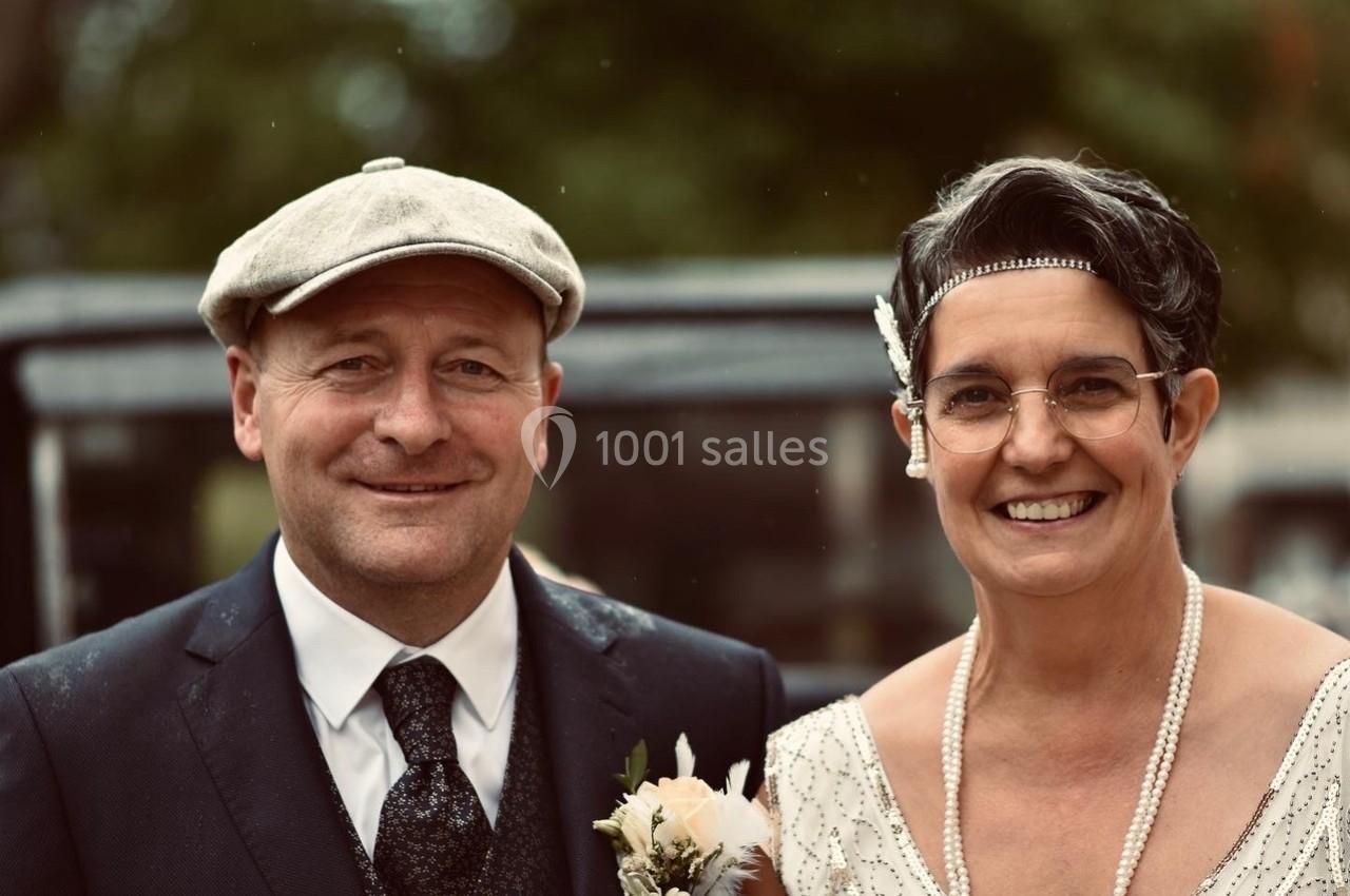 Un couple souriant en tenue élégante, l'homme portant une casquette et la femme un collier de perles.