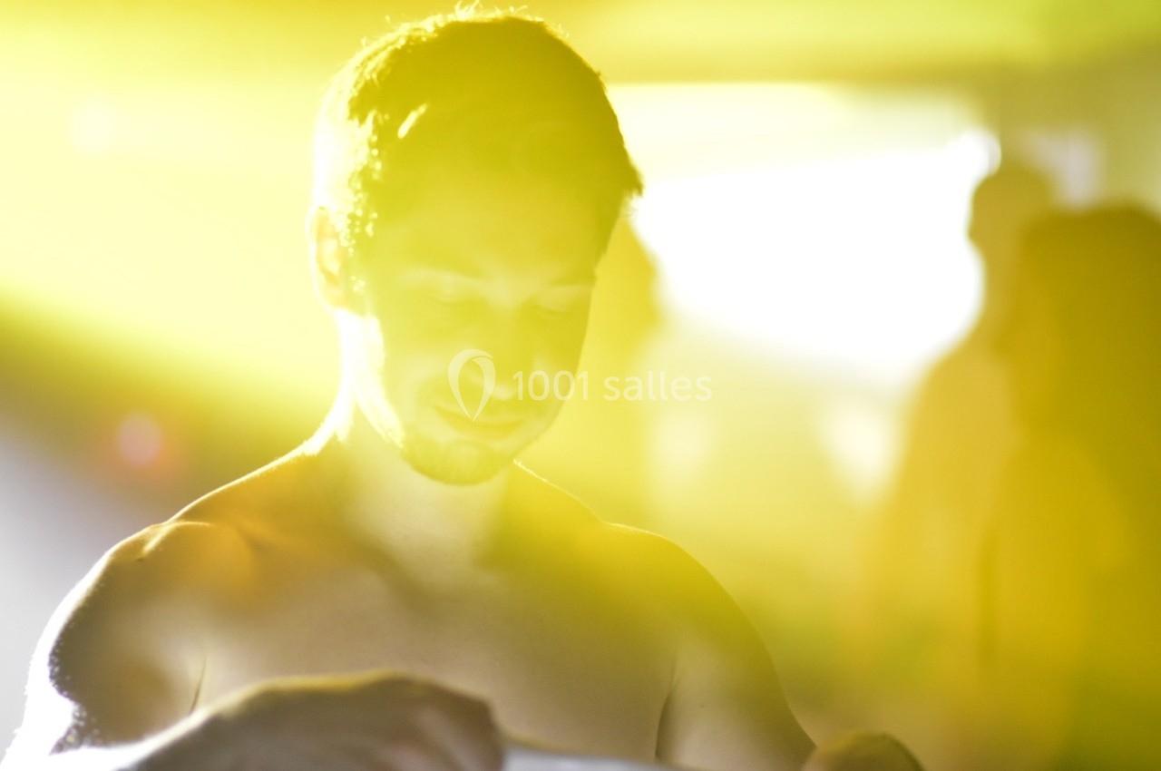 Un homme torse nu dans un environnement lumineux avec des silhouettes floues en arrière-plan.