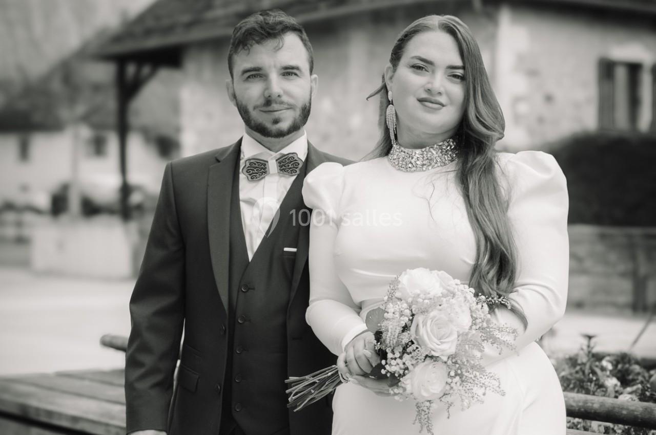 Un couple en tenue de mariage pose devant un bâtiment en pierre dans un cadre extérieur.