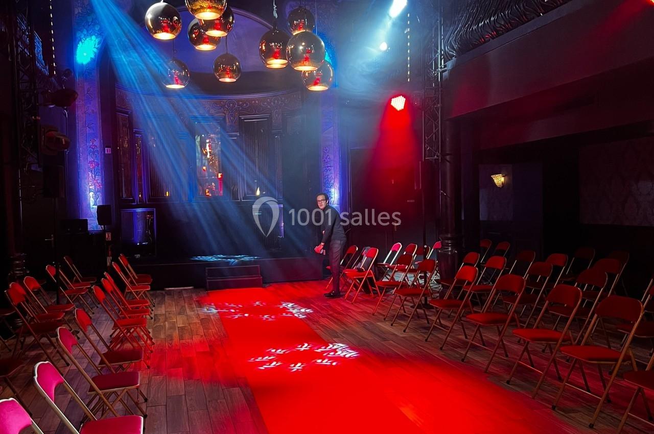 Salle de spectacle avec chaises pliantes alignées, éclairage coloré et projecteurs illuminant une scène.