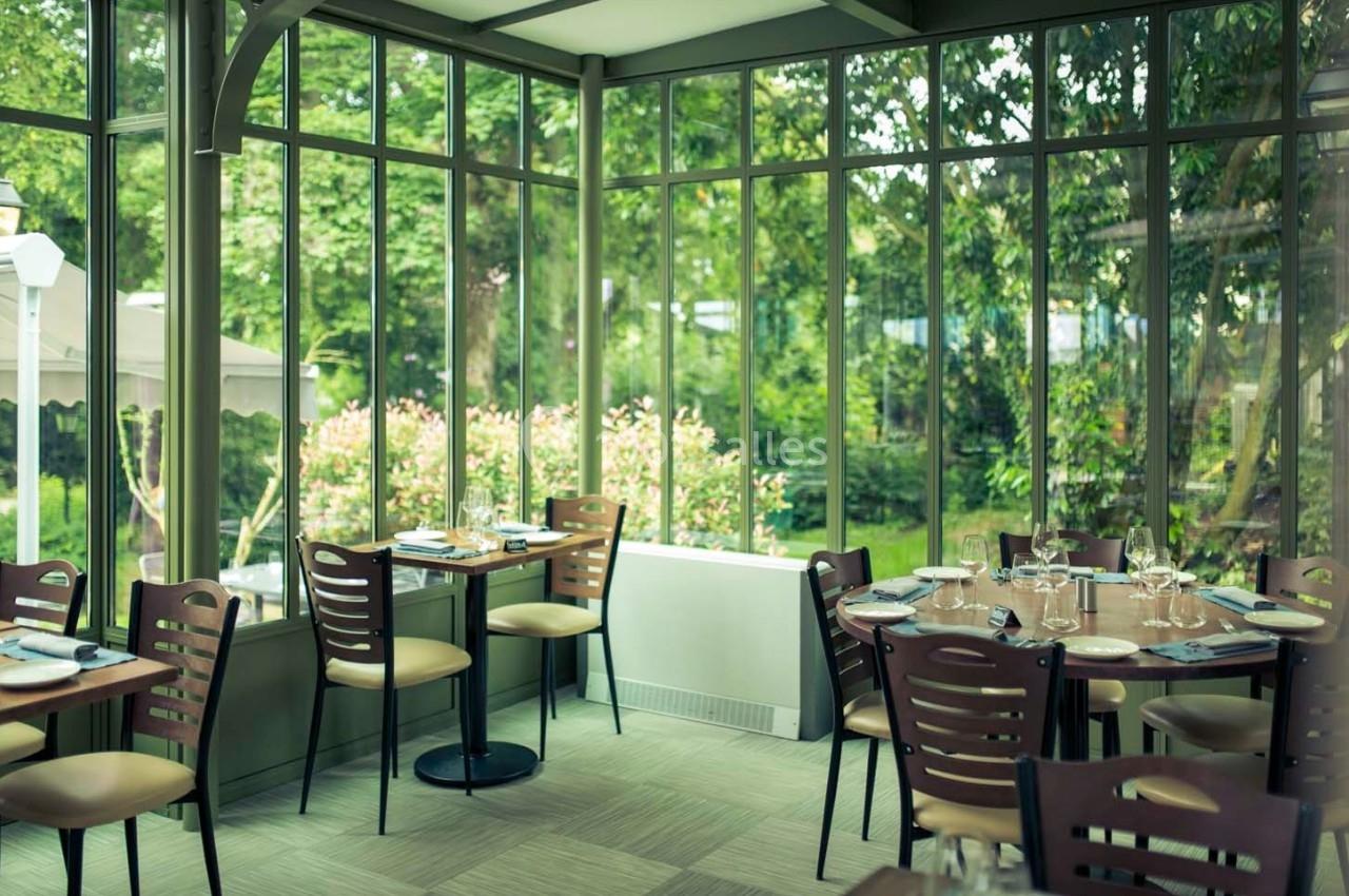 Salle de restaurant lumineuse avec baies vitrées donnant sur un jardin verdoyant, tables dressées pour le service.