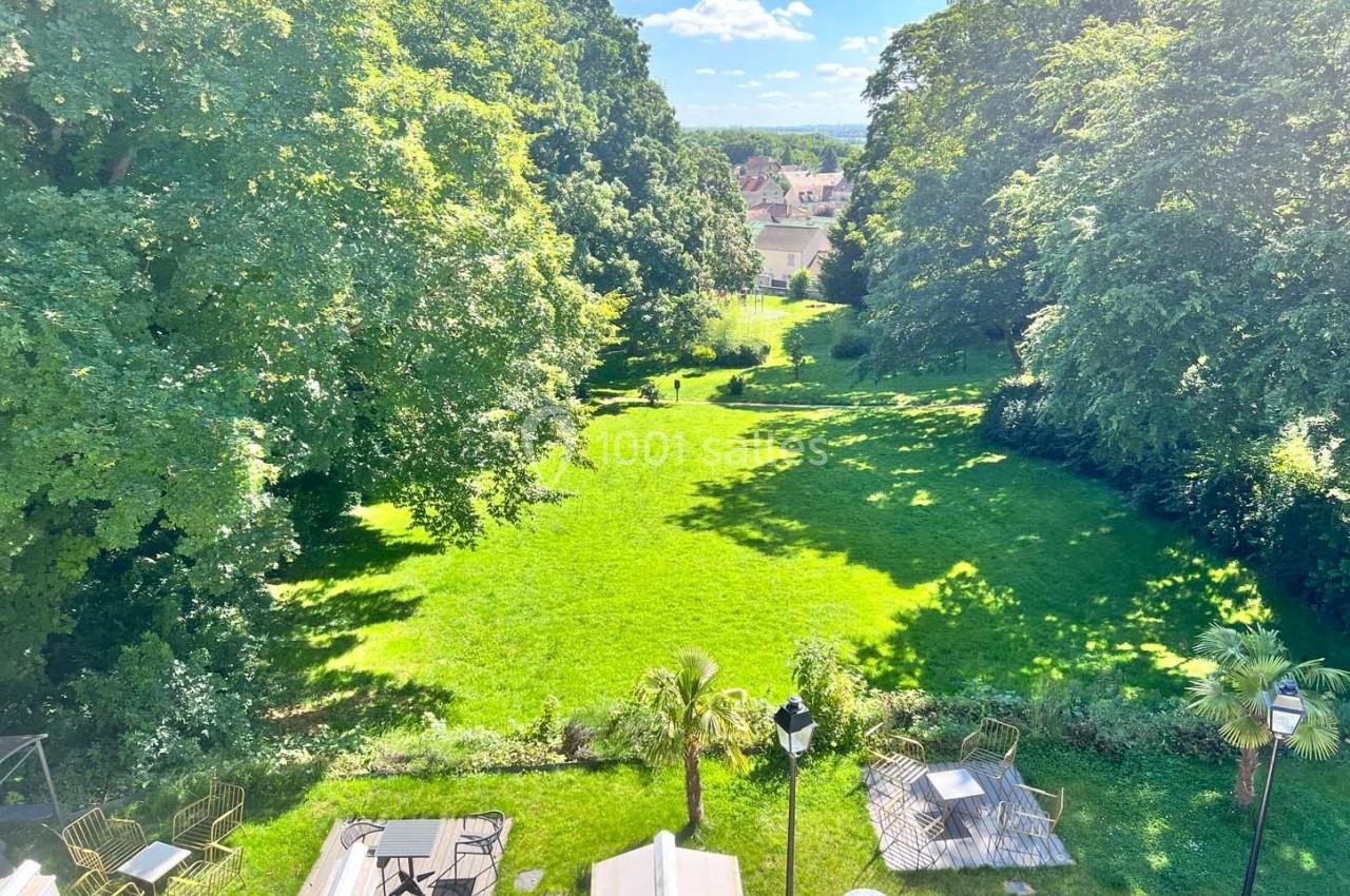 Vue d'un grand jardin verdoyant entouré d'arbres, avec quelques tables et chaises disposées en terrasse.