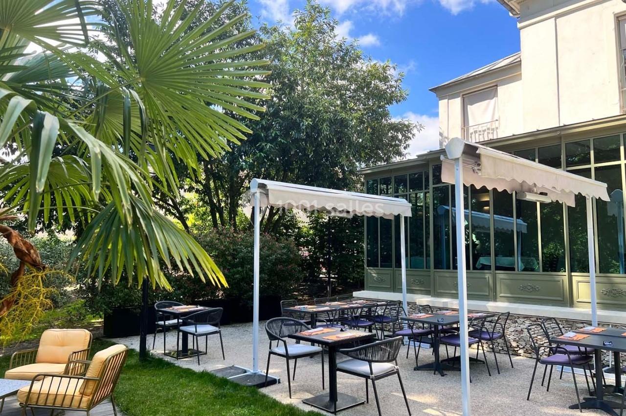 Terrasse extérieure avec tables et chaises, entourée de végétation, devant une maison avec grandes baies vitrées.
