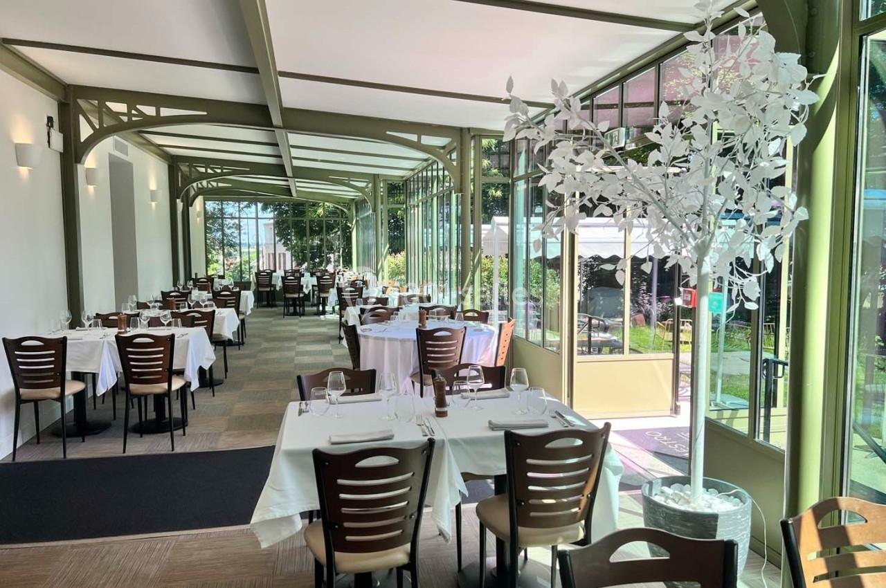 Salle de restaurant lumineuse avec grandes baies vitrées, tables dressées et vue sur un jardin extérieur verdoyant.