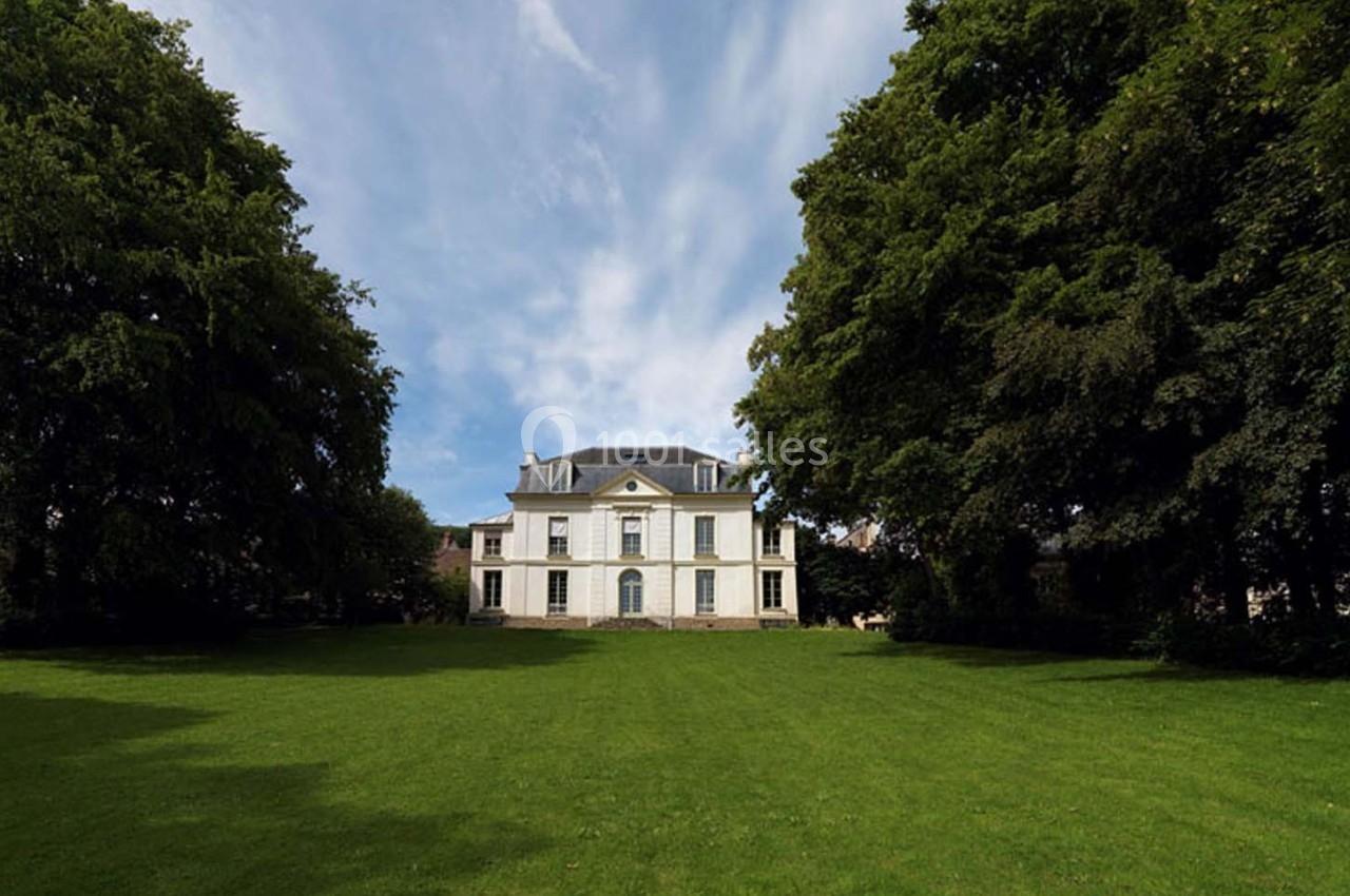 Manoir blanc entouré de grands arbres, avec une pelouse verte au premier plan et un ciel partiellement nuageux.