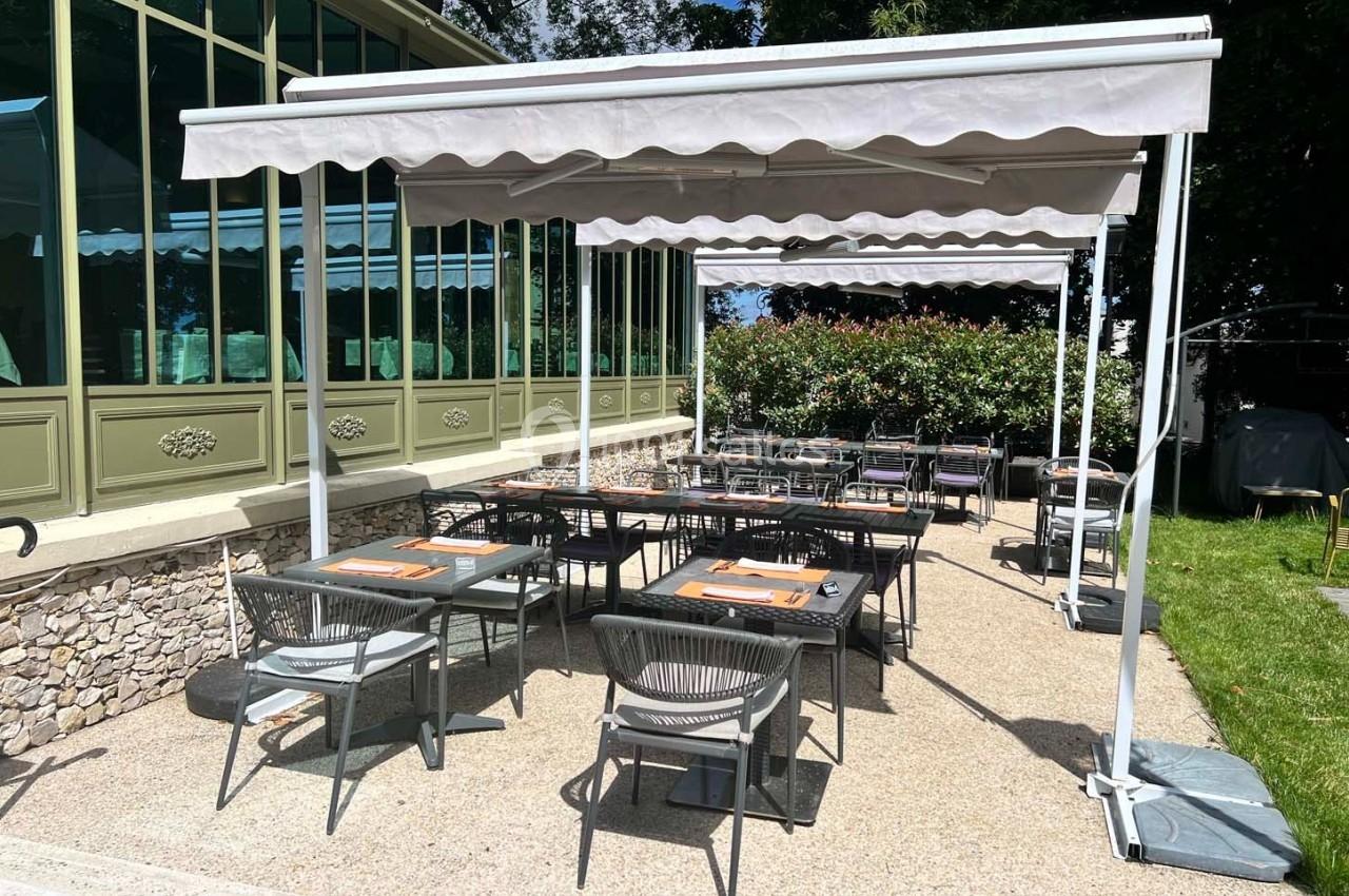 Terrasse extérieure d'un restaurant avec tables et chaises sous des stores, bordée de verdure et d'une façade vitrée.