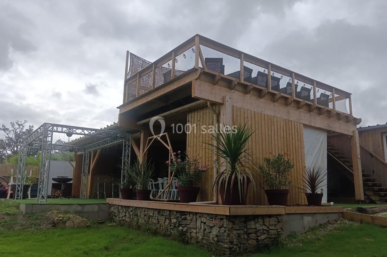 Structure en bois à deux niveaux avec terrasse supérieure, plantes en pots et ciel nuageux en arrière-plan.