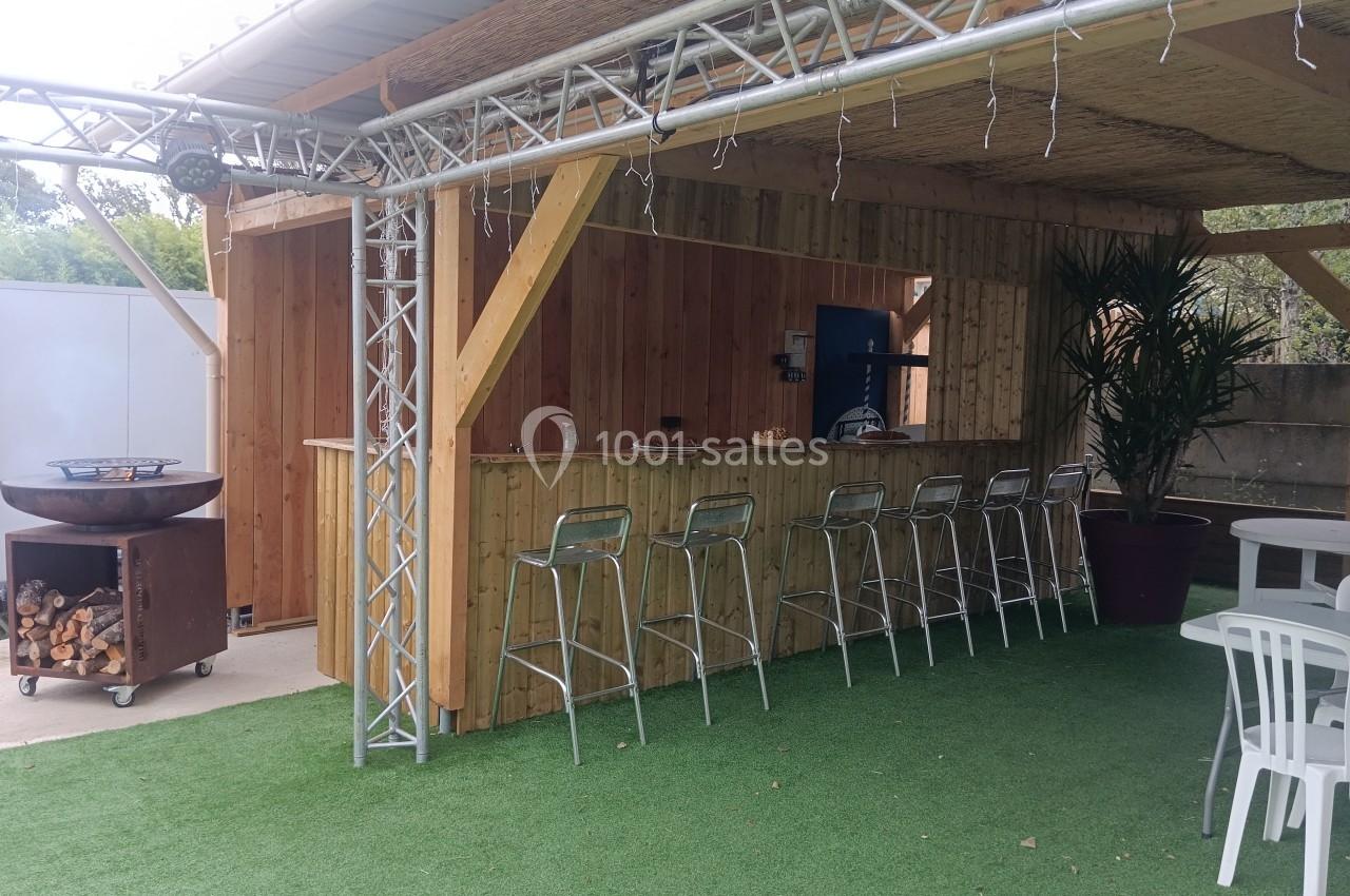 Bar extérieur en bois avec tabourets métalliques alignés, plancha à gauche et plantes en pot sur gazon synthétique.