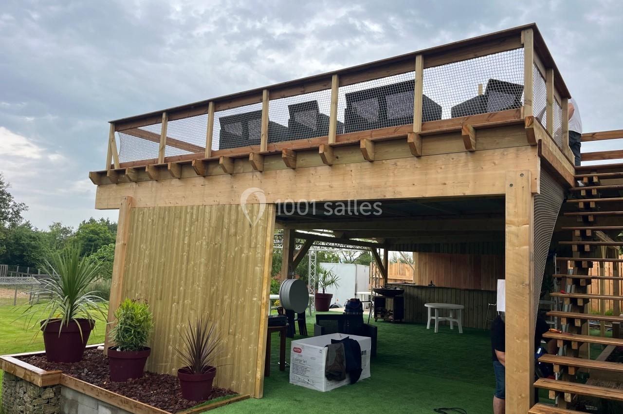 Structure en bois à deux niveaux avec terrasse aménagée en haut et espace couvert en bas, entourée de plantes.