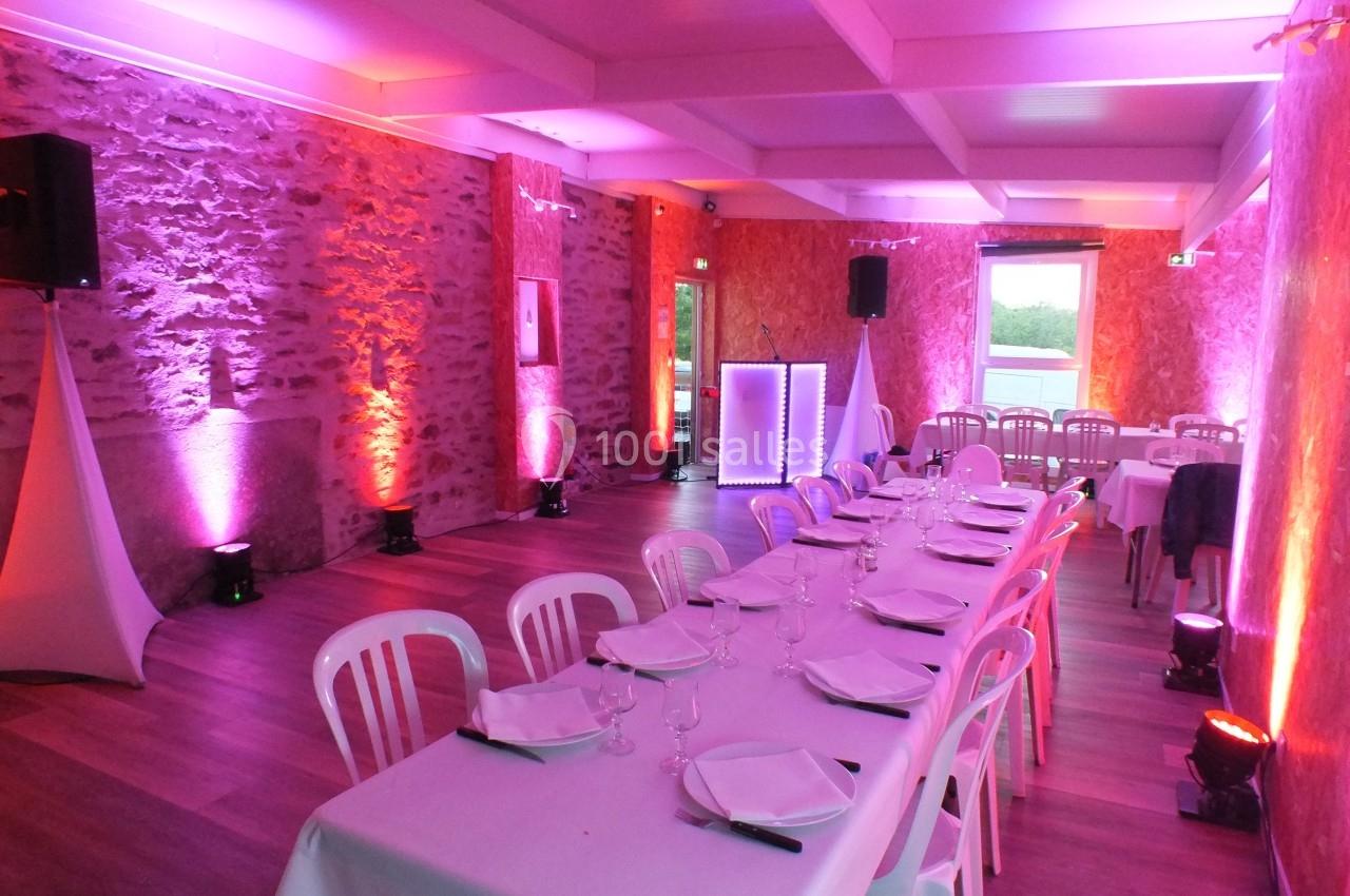 Salle de réception avec murs en pierre, éclairage coloré, tables dressées et chaises en plastique blanc.