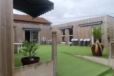 Cour extérieure avec sol en gazon synthétique, tables, chaises empilées et plantes en pots, entourée de bâtiments en bois.