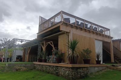 Cour extérieure avec sol en gazon synthétique, tables, chaises empilées et plantes en pots, entourée de bâtiments en bois.