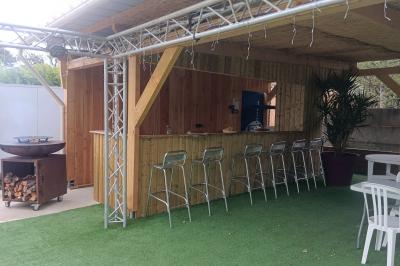 Cour extérieure avec sol en gazon synthétique, tables, chaises empilées et plantes en pots, entourée de bâtiments en bois.