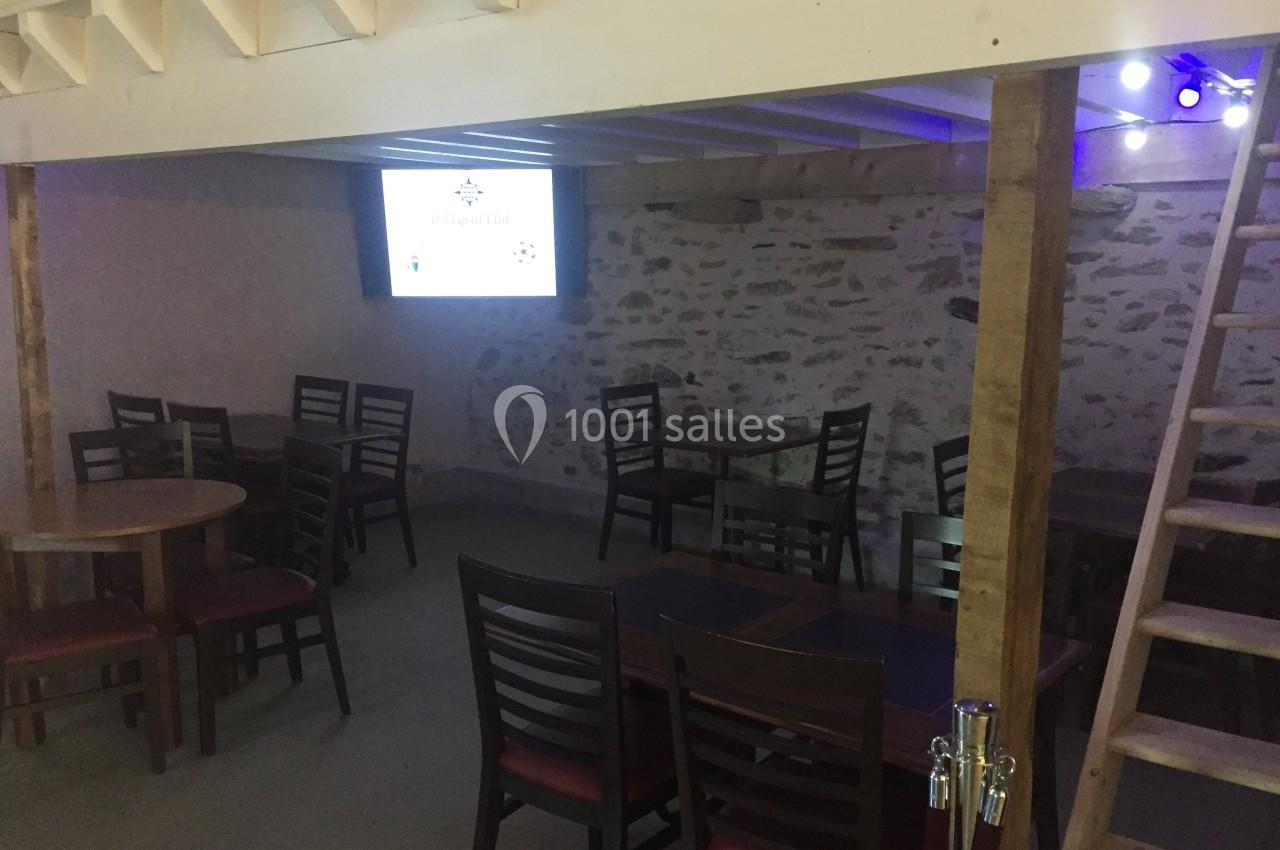 Salle avec des tables et chaises en bois, éclairage tamisé, mur en pierre apparente et écran allumé au fond.
