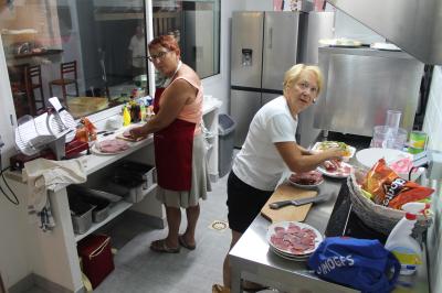 Cour extérieure avec sol en gazon synthétique, tables, chaises empilées et plantes en pots, entourée de bâtiments en bois.