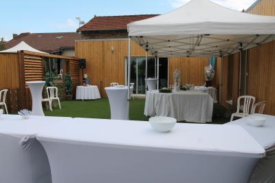 Cour extérieure avec sol en gazon synthétique, tables, chaises empilées et plantes en pots, entourée de bâtiments en bois.