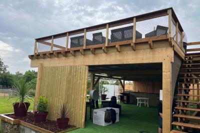 Cour extérieure avec sol en gazon synthétique, tables, chaises empilées et plantes en pots, entourée de bâtiments en bois.