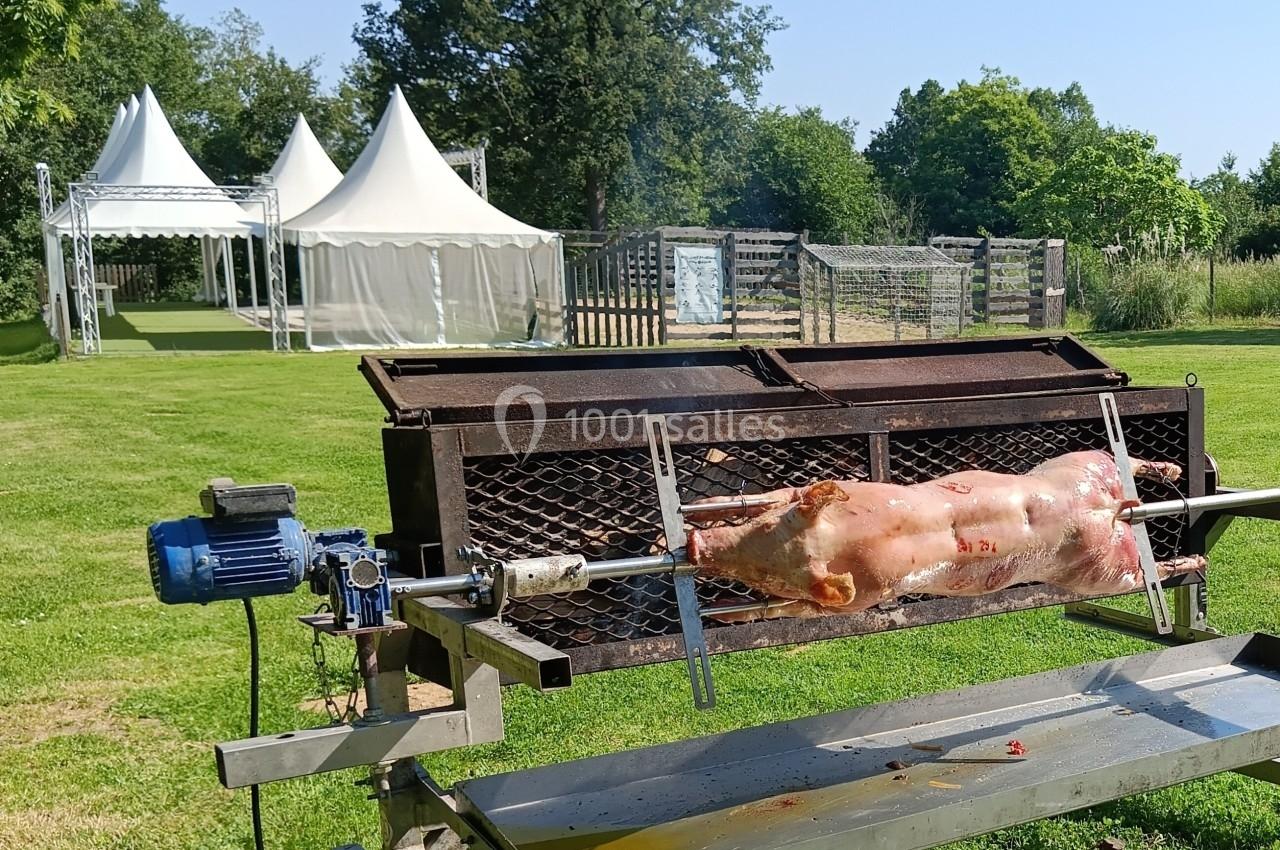 Un cochon rôti à la broche en plein air, avec des tentes blanches et un espace vert en arrière-plan.