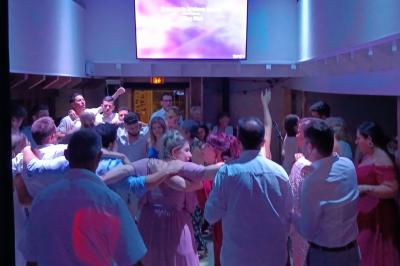 Cour extérieure avec sol en gazon synthétique, tables, chaises empilées et plantes en pots, entourée de bâtiments en bois.