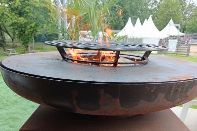 Cour extérieure avec sol en gazon synthétique, tables, chaises empilées et plantes en pots, entourée de bâtiments en bois.