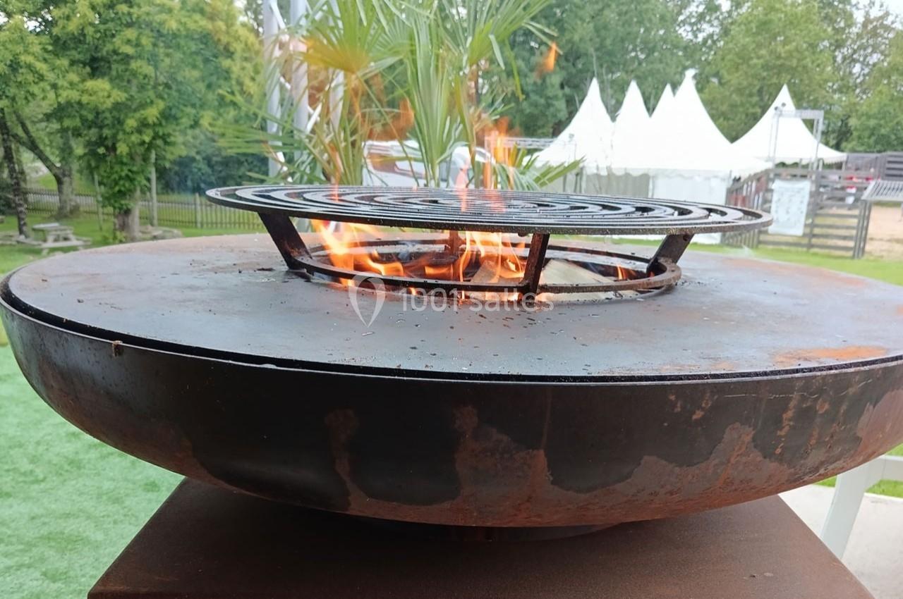 Barbecue circulaire en métal avec des flammes visibles, installé en extérieur près de végétation et de tentes blanches.