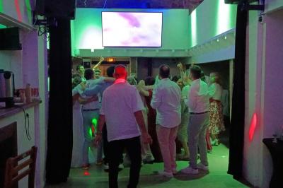 Cour extérieure avec sol en gazon synthétique, tables, chaises empilées et plantes en pots, entourée de bâtiments en bois.