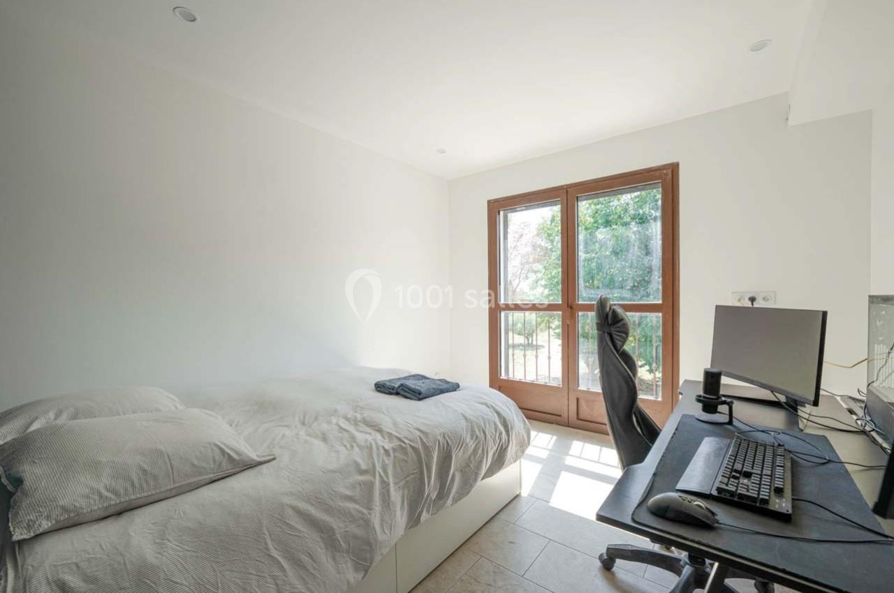 Chambre lumineuse avec lit double, bureau équipé d'un ordinateur et grande fenêtre donnant sur un espace extérieur verdoyant.