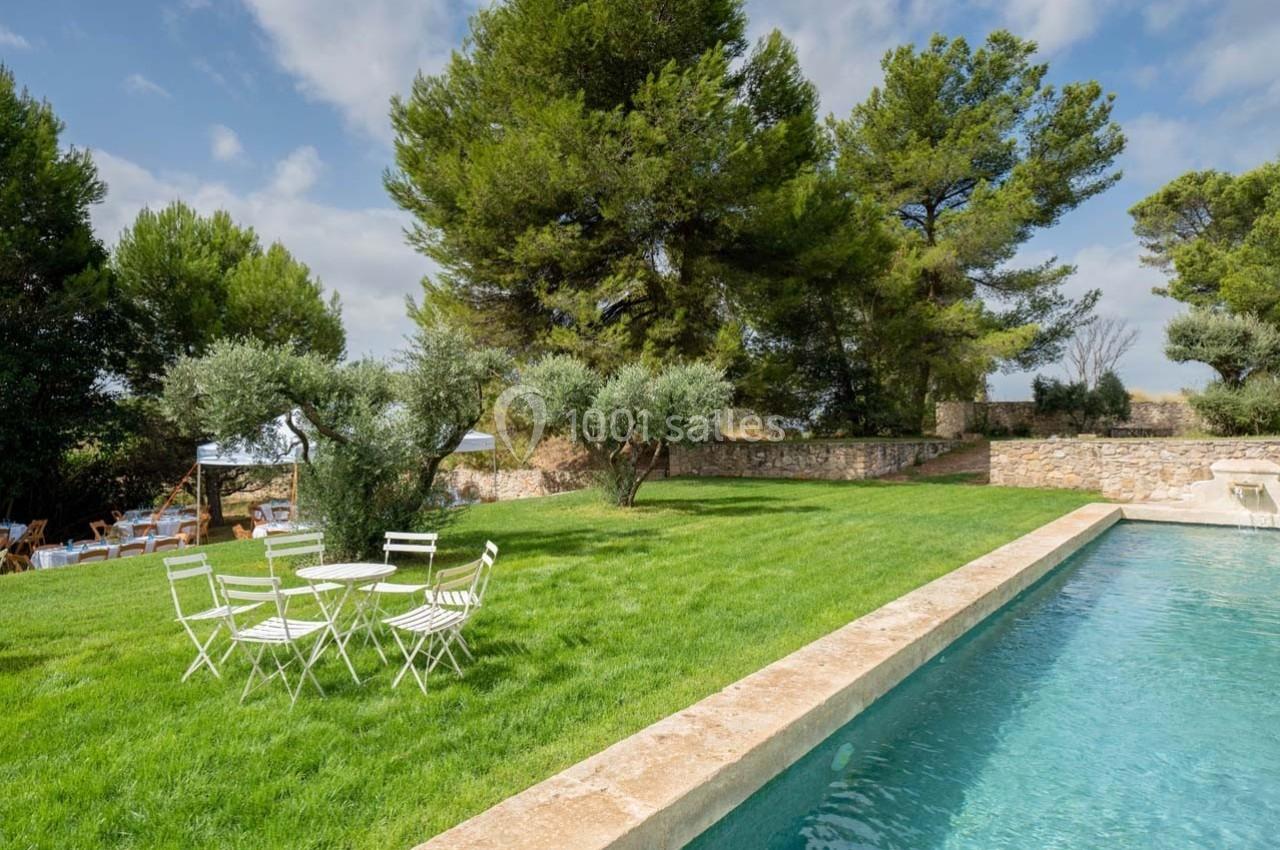 Jardin avec pelouse, piscine rectangulaire, table et chaises blanches, entouré d'arbres et d'un mur en pierre.