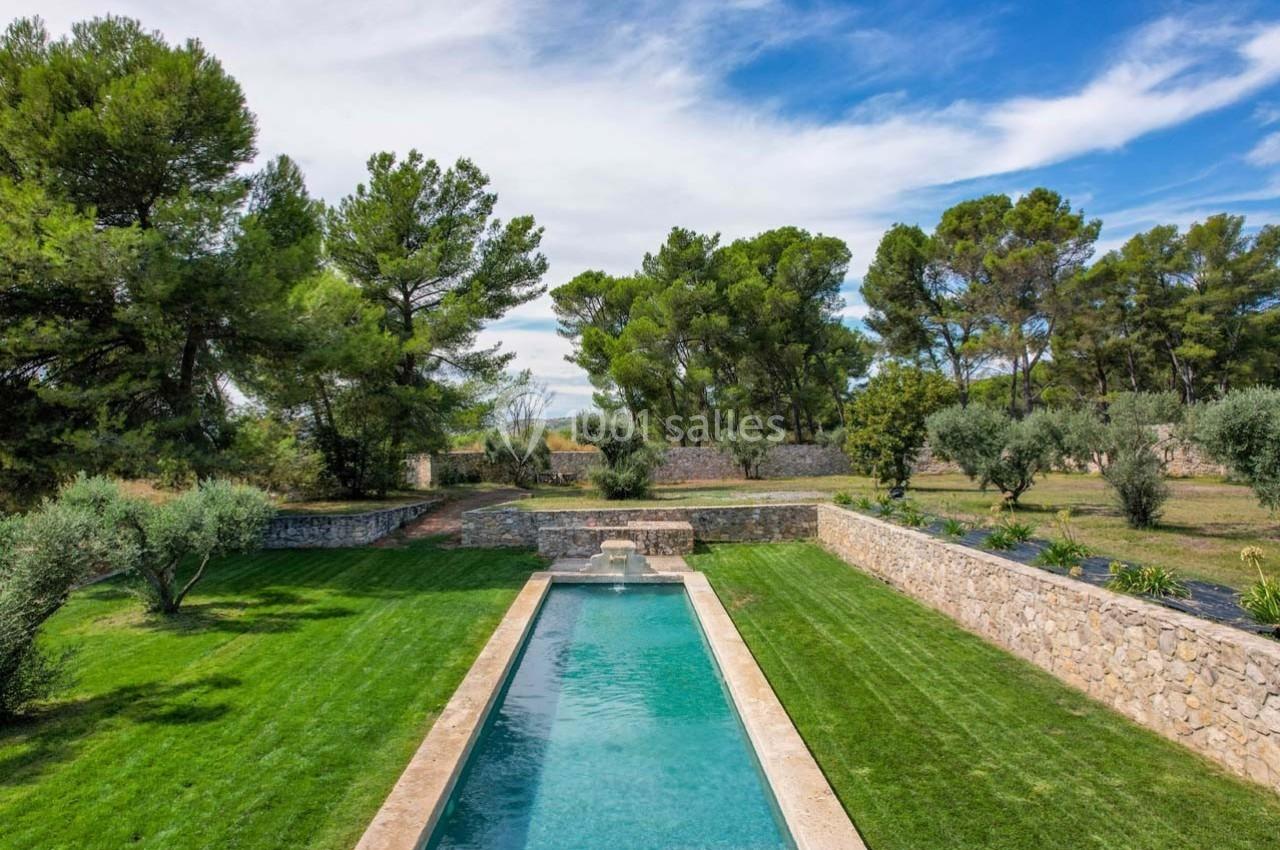 Piscine rectangulaire entourée de pelouses, d'arbres et de murets en pierre sous un ciel partiellement nuageux.