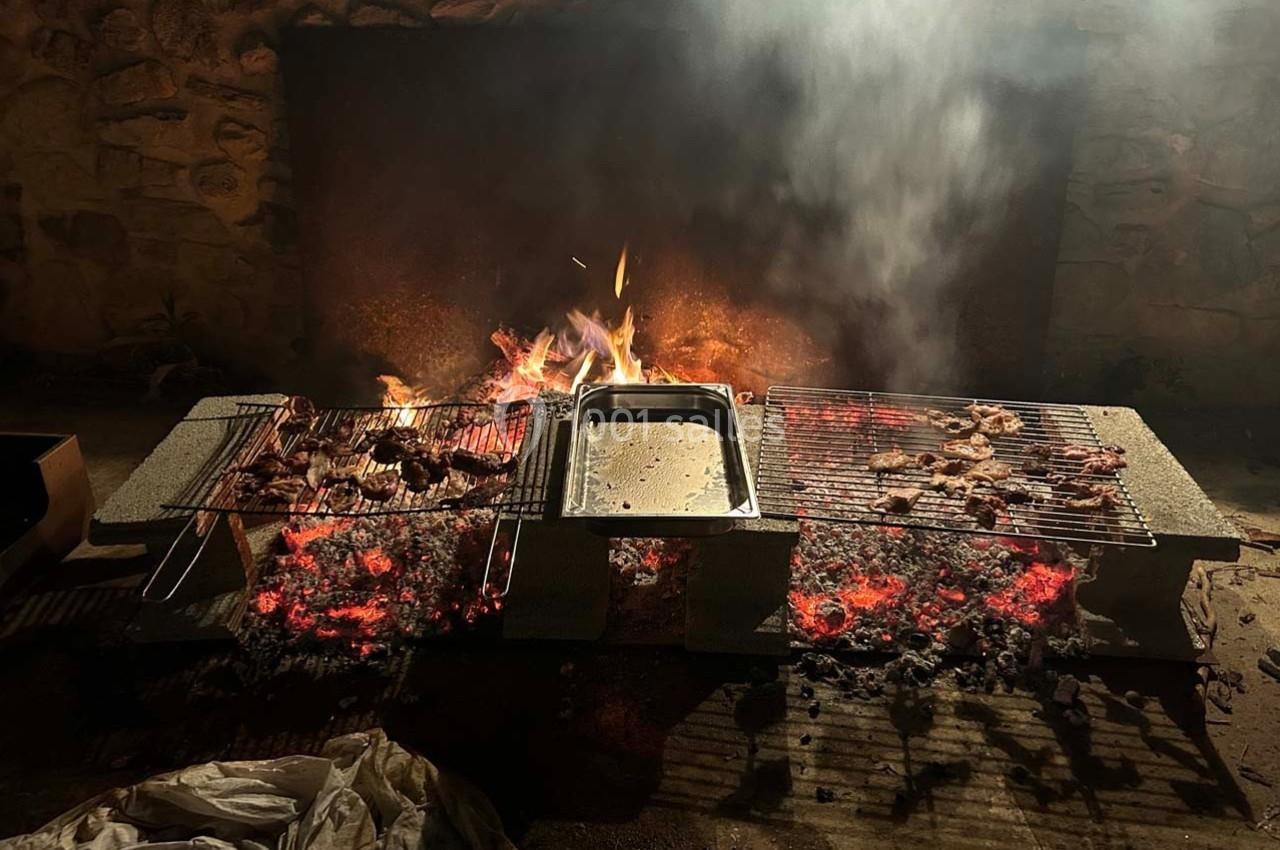 Grillades de viande sur des grilles au-dessus de braises rouges, avec un feu actif en arrière-plan.