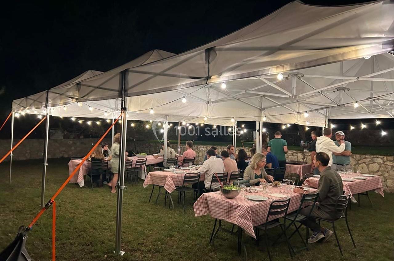 Des personnes dînent sous des tentes éclairées par des guirlandes lumineuses, installées sur une pelouse en soirée.