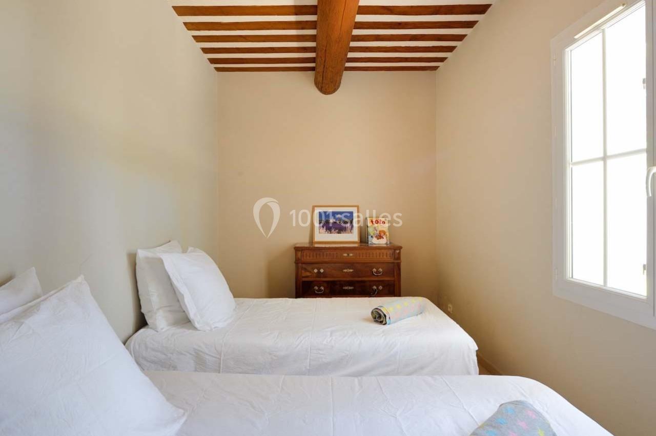 Deux lits simples avec draps blancs dans une chambre lumineuse au plafond en poutres apparentes, avec une commode en bois.