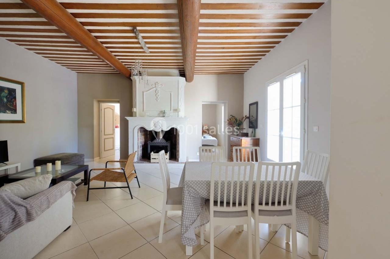 Salle à manger lumineuse avec table, chaises blanches, cheminée décorative et poutres apparentes au plafond.