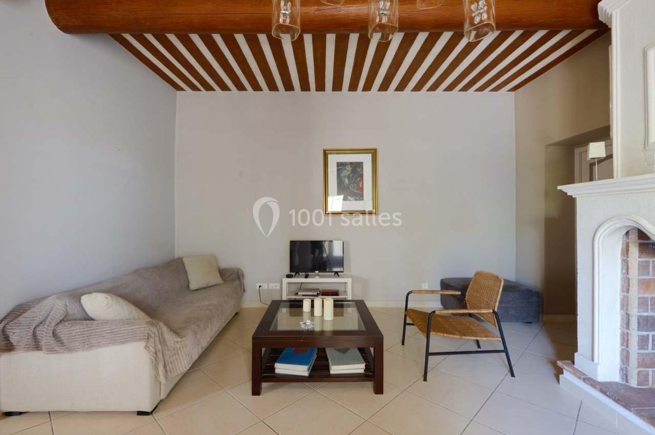 Salon avec canapé beige, table basse en bois, fauteuil en osier, télévision et cheminée en briques.