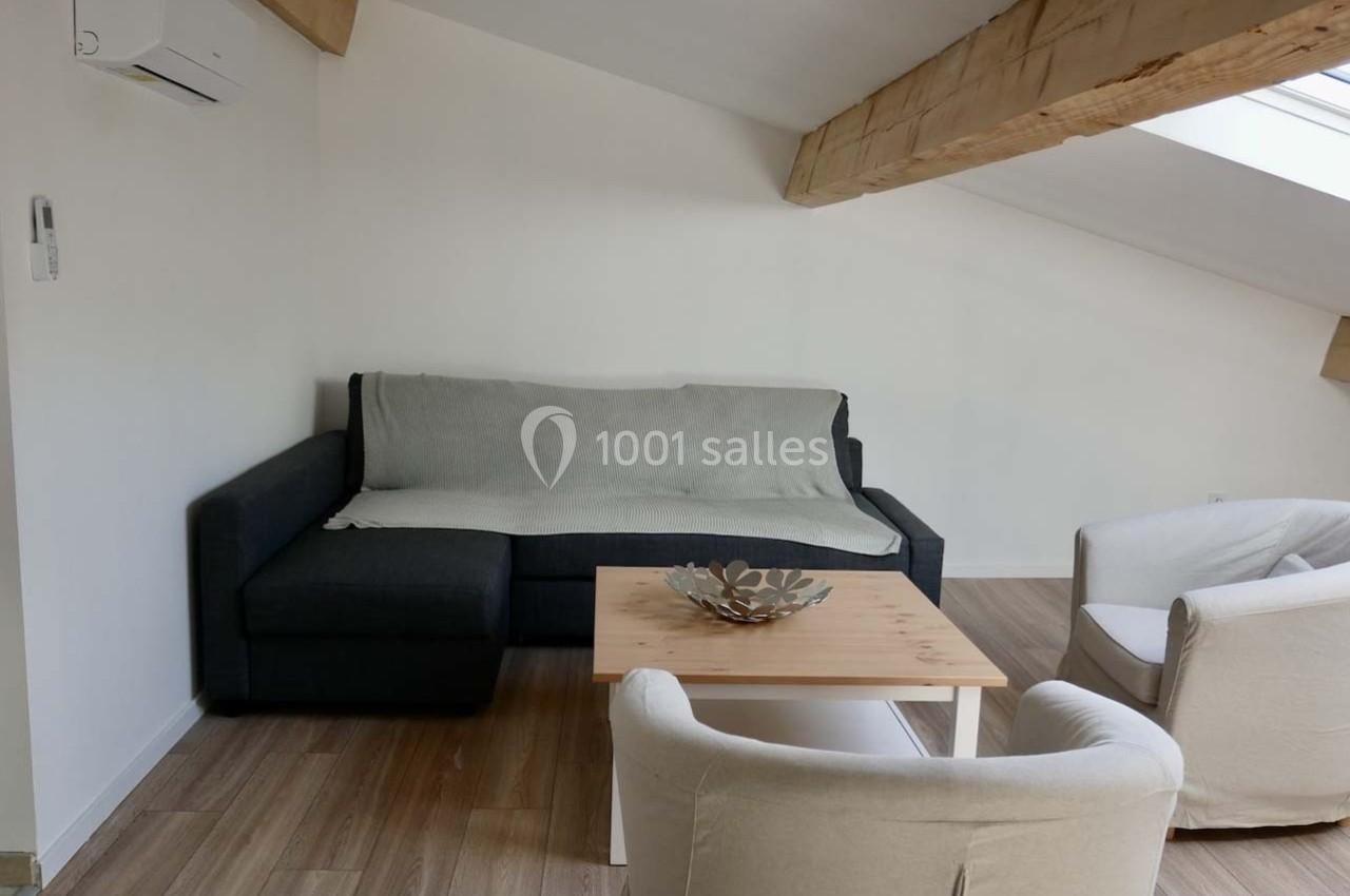 Salon mansardé avec canapé d'angle gris, table basse en bois, deux fauteuils blancs et poutres apparentes.