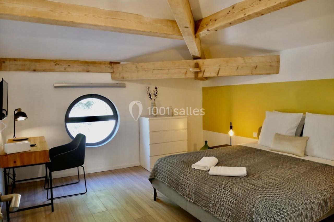 Chambre lumineuse avec poutres apparentes, lit double, bureau, commode blanche et fenêtre ronde.