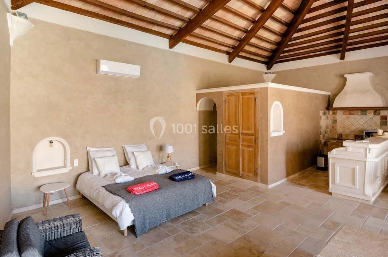 Chambre lumineuse avec lit double, kitchenette, plafond en bois et murs beige, dans un style rustique.