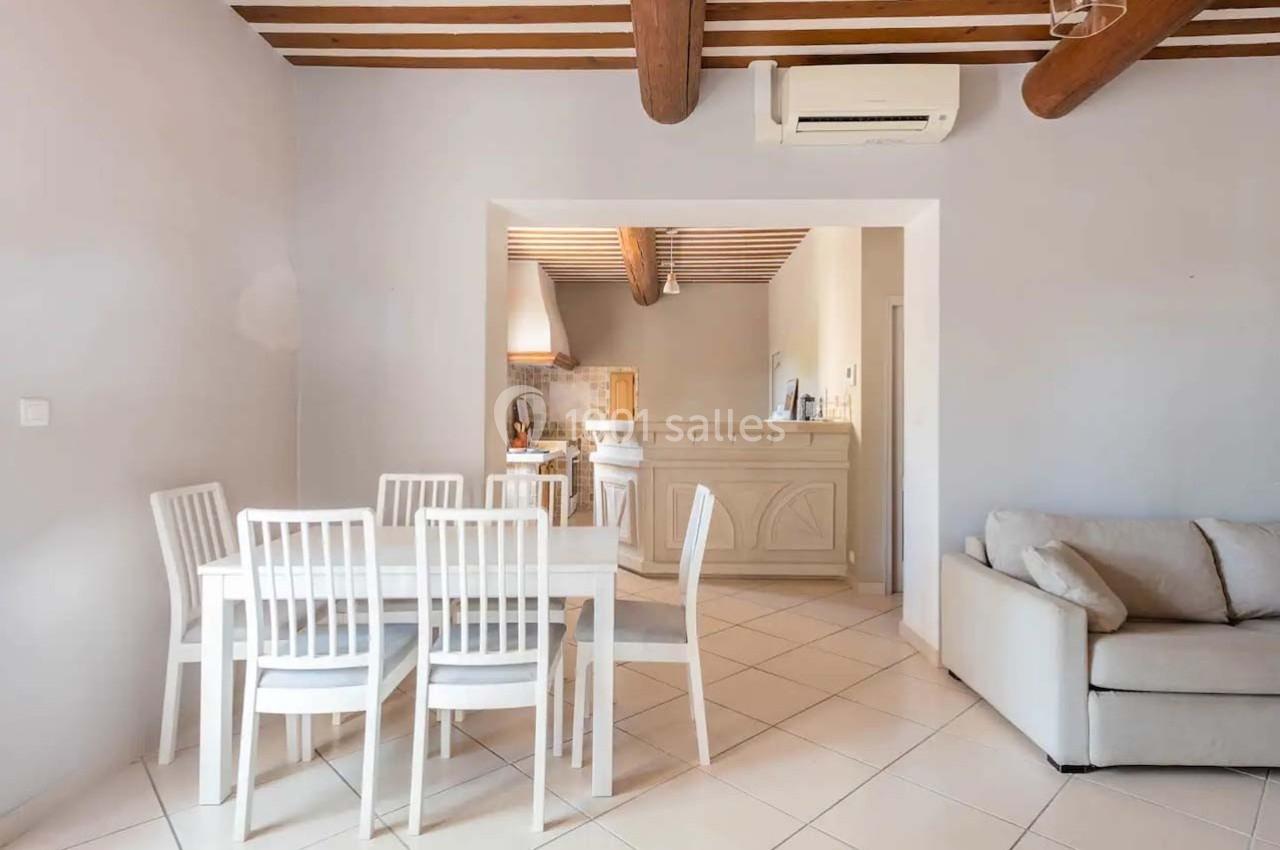 Salle à manger lumineuse avec table blanche et chaises, ouverte sur une cuisine équipée avec poutres apparentes au plafond.