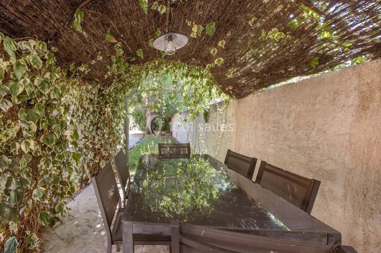 Table en verre entourée de chaises sous une pergola en branches avec des plantes grimpantes et un éclairage suspendu.