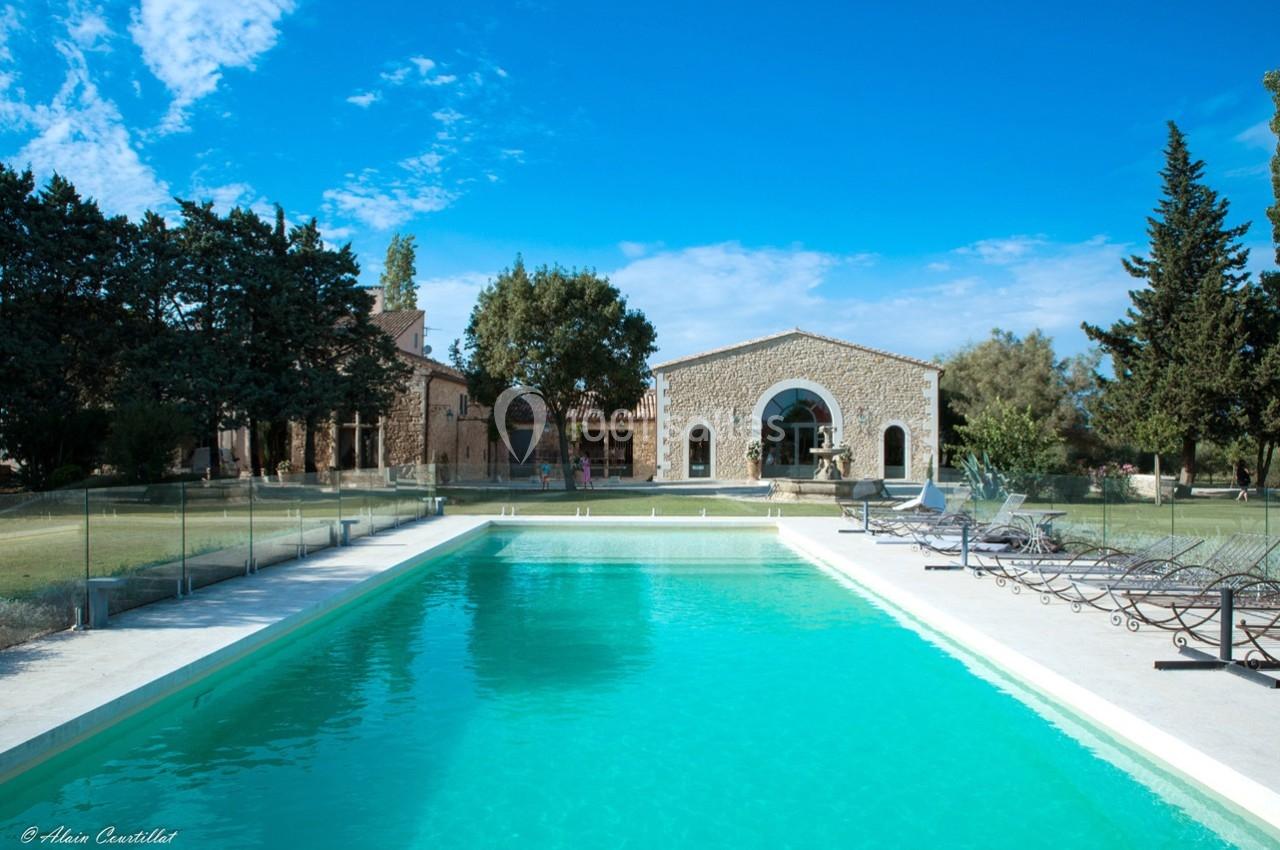 Piscine extérieure entourée de transats, devant une maison en pierre avec jardin arboré sous un ciel bleu.