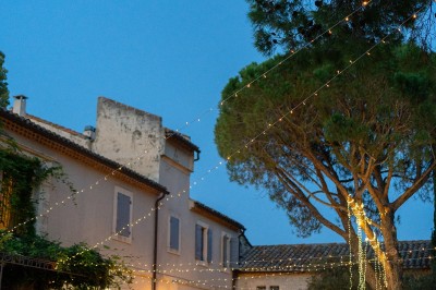 Tables dressées pour un dîner en plein air sous des guirlandes lumineuses, devant un bâtiment ancien au crépuscule.