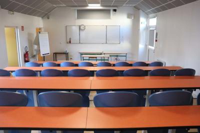 Salle de classe vide avec des rangées de tables et chaises, tableau blanc et écran au mur.
