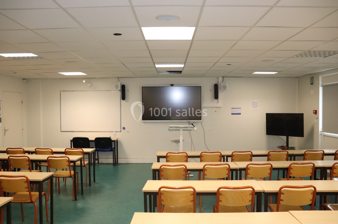 Salle de classe vide avec des rangées de tables et chaises, tableau blanc, écran interactif et éclairage au plafond.