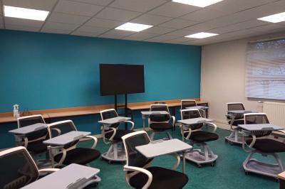 Salle de classe vide avec des rangées de tables et chaises, tableau blanc et écran au mur.